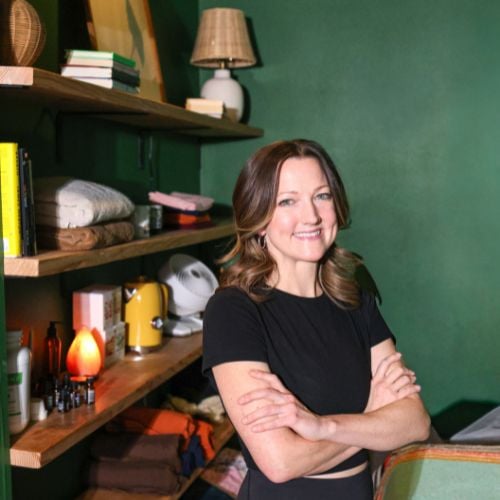 Woman in black t-shirt standing with arms crossed before wooden shelves against green wall