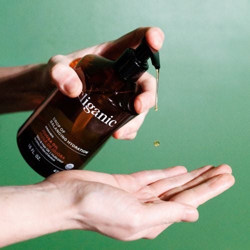 Hand dispensing organic skincare serum from dark brown bottle onto palm against green background